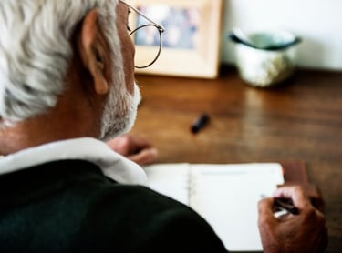 Senior man with white hair and glasses writing notes in a paper notebook at a wooden desk.