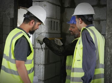 Construction workers in hard hats and safety vests measure a concrete block wall on a building site.