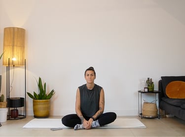 A woman sits in a cross-legged yoga pose on a white mat in a minimalist home studio.