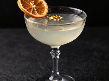 An elegant, professional close-up photograph of a craft cocktail in a crystal coupe glass, garnish of dried orange and gold flakes, dramatic side lighting, South American styling, dark editorial backdrop.