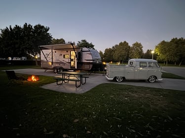 A Jayco travel trailer parked at a campsite next to a vintage Volkswagen truck and campfire at dusk.