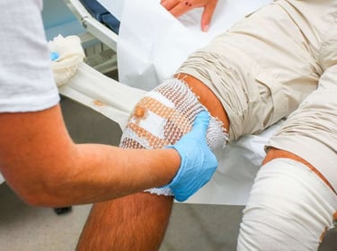 A healthcare professional in blue gloves applies a sterile gauze bandage to a patient's injured knee.