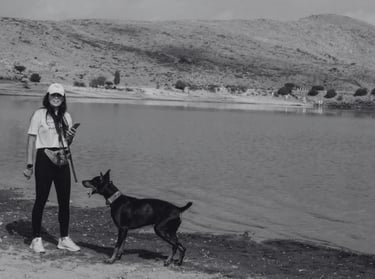 Una mujer junto a su Doberman Pinscher negro en la orilla de un lago con colinas al fondo.