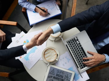 Business professionals shaking hands over a table with a laptop, digital marketing reports, and coffee.