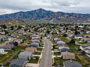 Aerial view of Riverton Utah neighborhood, curving residential streets, Wasatch peaks in background, overcast daylight, green parks and modern homes visible