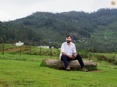 Traveler sitting on a tree trunk at Mannavanur Sheep Farm, surrounded by rolling meadows and hills.