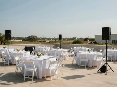 Wide shot of a neatly staged outdoor event area showing tables, chairs, and a sound speaker together in one frame on an open property, clean noon light, no attendees — the complete setup as the subject