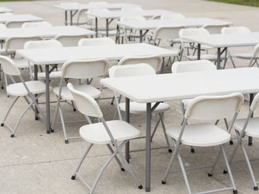Rows of white folding chairs and rectangular trestle tables arranged in formation on a clean concrete surface, neutral daylight, wide environmental framing showing the full setup
