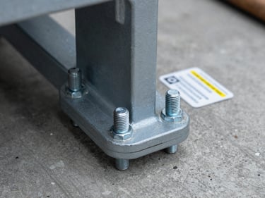 Extreme close-up of a structural module connection point — heavy-gauge bolt, load plate and certification tag in sharp focus, gray concrete floor below, shallow depth of field, overcast industrial light