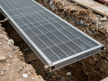 Overhead close-up of a modular anti-slip walkway panel spanning a deep excavation trench, steel grating texture in sharp focus, construction debris visible below, overcast daylight