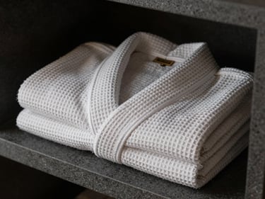 Close-up of a spa robe folded on a dark grey stone shelf, studio strobe lighting picking out the waffle-weave texture and a woven label on the collar, no people, tight framing