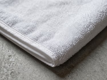 Extreme close-up of a hotel hand towel corner on a grey concrete surface, tight studio strobe lighting revealing the looped terry weave texture and a faint embroidered border detail, no people, overhead angle