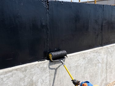 Worker applying liquid black bitumen coating to a concrete retaining wall with a roller, high contrast daylight, architectural construction details