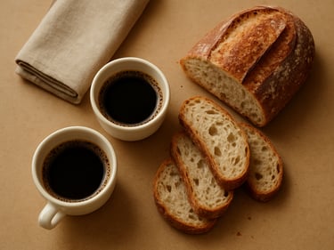 Close-up overhead of artisanal coffee-break spread — small cups, sliced pão artesanal and a folded linen napkin on a warm beige surface, soft diffused daylight, no people