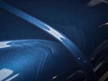 Close-up macro of automotive paint under studio spot lighting, swirl marks and fine scratches clearly visible across dark blue metallic surface before correction treatment, controlled directional light at low angle revealing depth of surface defects
