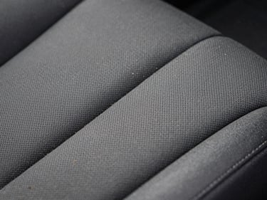 Close-up of automotive interior upholstery texture under controlled diffused lighting, fabric weave detail visible with embedded dirt particles before extraction treatment, high contrast revealing surface condition