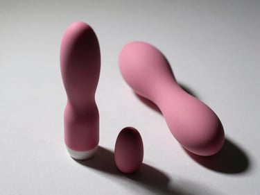 Close-up straight-on studio shot of two vibrators on a white-grey surface — one compact bullet vibrator and one larger curved model — directional side lighting revealing silicone texture and material detail, neutral background, no people, no lifestyle props.