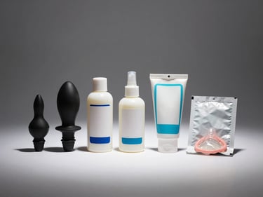 Straight-on studio shot of several accessory items — anal plug, lubricant bottle, and condom pack — arranged in a clean row on white surface, strong directional overhead lighting, clear label-free product forms, neutral background, no people
