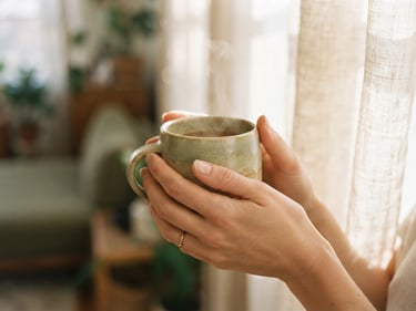 Soft focus close-up of hands gently holding a warm ceramic mug, natural light filtering through a sheer curtain in the background, muted sage and ecru tones, shallow depth of field, warm editorial photography.