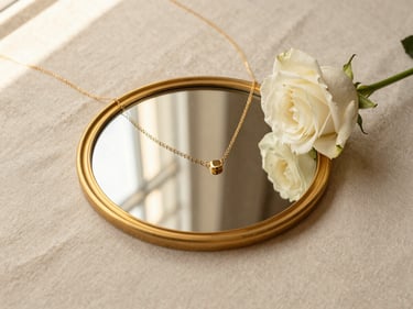 An editorial flat-lay of a gold-rimmed mirror, a delicate gold necklace, and a fresh white rose on a cream linen background, warm window reflections.