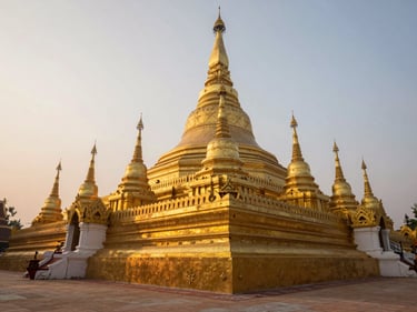 Close-up of Pha That Luang golden stupa in Vientiane at dawn, warm haze diffusing morning light across ornate gilded spires, low angle looking upward, stone courtyard in foreground