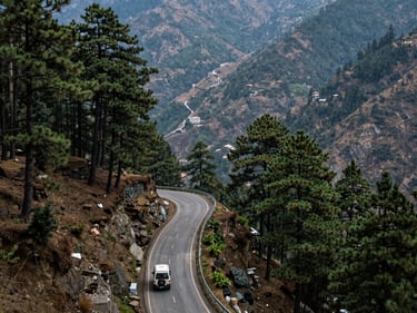 Wide shot of a mountain road in Uttarakhand with a vehicle winding through dense pine forest, natural daylight, valley visible below