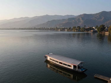 Wide environmental shot of Dal Lake in Kashmir from a high vantage point, a houseboat visible at the water's edge, mountains in background, morning light