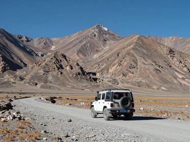Wide shot of Leh-Ladakh high-altitude plateau with a 4x4 vehicle on a gravel road, barren mountains in the background, sharp midday sunlight