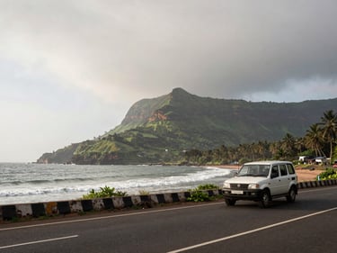 Wide environmental shot of a coastal Maharashtra road with a vehicle in the foreground, the Western Ghats visible in the background, overcast morning light