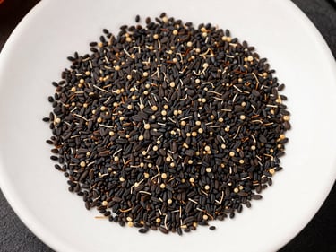 Overhead flat-lay of dried black seed (nigella sativa) pods and loose seeds scattered on a white ceramic tray under north-facing studio light, seed oil dropper bottle visible at the edge