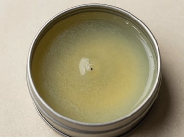Extreme close-up overhead studio shot of a flat open tin of herbal lip balm on a neutral parchment surface, waxy surface texture and faint green-amber colour visible, bright studio daylight, no lifestyle props