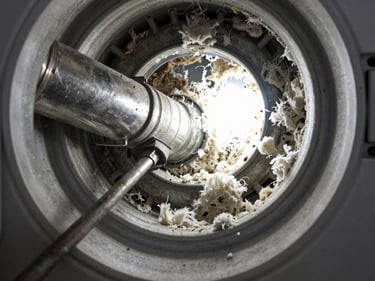 Interior view of a clogged dryer vent pipe, dense lint blockage visible under bright work light, metal duct walls showing buildup, technician tool entering from the left