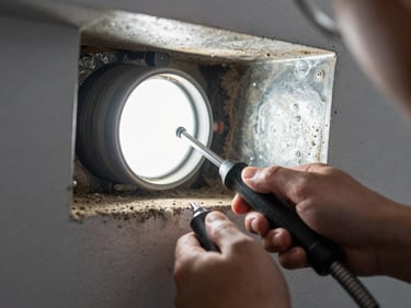 Close-up of technician hands using a flexible camera probe inside a residential air duct, bright LED illuminating dust-caked metal walls, tool grip visible in foreground