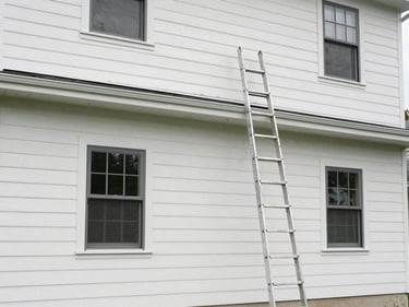 Wide shot of a two-story home exterior freshly painted in natural daylight, clean siding lines, new trim color visible, ladder resting against wall, no people