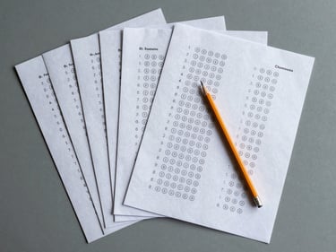 Flat-lay overhead of B1 exam practice papers fanned out on a grey surface, a pencil placed diagonally across them, one sheet showing multiple-choice answer bubbles, cool natural light, no text legible, muted tones