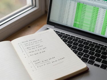 Overhead view of a notebook with handwritten financial model notes next to a laptop showing Excel rows with formula cells highlighted in green, daylight from a window
