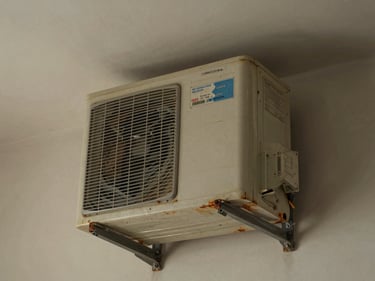 Old window AC unit wedged into a wall opening in a Noida flat, natural indoor light, visible rust on the casing edges, unit label partially faded
