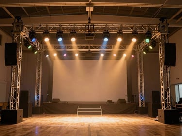 In-situ sound and lighting rig at an indoor event venue — truss structure with coloured LED par cans and speakers visible, warm amber wash across a decorated stage behind, no people, wide environmental shot from floor level