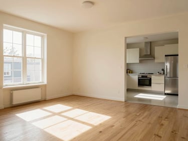 Wide shot of a sunlit two-bedroom apartment living room, cream walls, open-plan kitchen visible in background, natural window light from the left