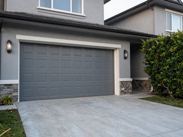 Wide shot of a residential property exterior with a double garage door closed, photographed in natural morning light, grey rendered walls, concrete forecourt, mature hedge to the right, no people