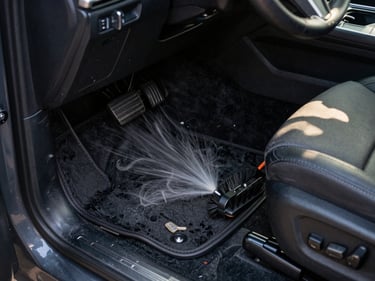 Overhead close-up of a technician vacuuming the floor mat of a dark SUV interior, natural morning light coming through the open passenger door, upholstery brush resting on the seat nearby