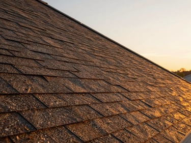 Close-up of a steep-pitch residential roofline at golden hour, new architectural shingles installed flush, shadow lines showing precise course alignment, warm amber light raking across the surface