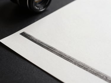 Overhead close-up of a fine-art paper sheet under controlled north-facing daylight, smooth bright-white surface showing ink density at the edge of a printed motif, dark studio surround