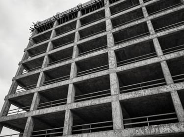 Wide architectural shot looking up the facade of a concrete mid-rise under construction in an urban Pacific Northwest setting, steel rebar grid visible through open floor plates, overcast diffused daylight, stark high-contrast monochrome
