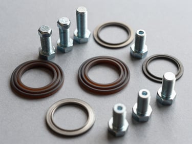 Overhead macro of spiral-wound gaskets and hex bolt sets arranged by size on a flat grey surface, studio lighting revealing metallic finish and PTFE windings in precise detail