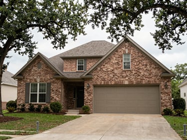Tight exterior shot of a brick ranch-style home in a Houston suburb, wide driveway, two-car garage door, established oak trees framing the facade, bright overcast documentary light, no people