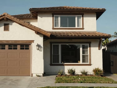 Close documentary shot of a craftsman-style home exterior in a Los Angeles residential neighborhood, stucco facade with wood trim, a garage door visible on the left, clear afternoon daylight, no people