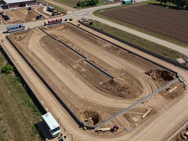 Aerial view of a land development site with surveyed lots and foundation work underway, construction equipment parked at plot boundaries, a command trailer visible at the site edge, clear midday light casting hard shadows across the graded earth