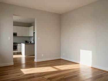 Corner-angle interior shot of a one-bedroom apartment, natural daylight across hardwood floors, open kitchen visible in background, empty and clean, architectural framing