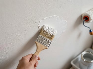 Overhead shot of a painter's brush loaded with warm white paint sweeping across a textured interior wall, soft diffused daylight, paint tin and roller tray visible at the edge of the frame